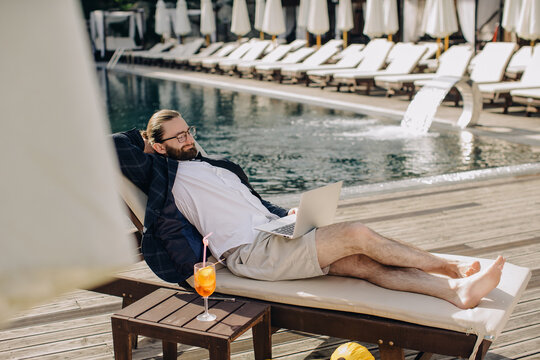 Businessman relaxing by pool working on laptop with cocktail