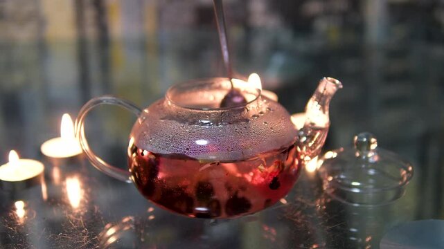 and stir red hibiscus tea with a spoon in a glass teapot on a dark background by candlelight. girl preparing for a tea ceremony. Lights a candle and incense. On a low wooden table with tea 