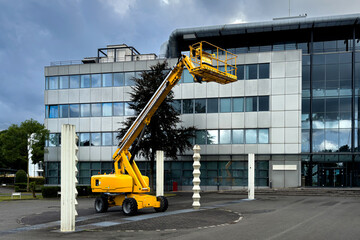 A bright yellow lift is raised near a contemporary office structure, showcasing its height and work capability. Dark clouds loom over the location, suggesting an impending change in weather.