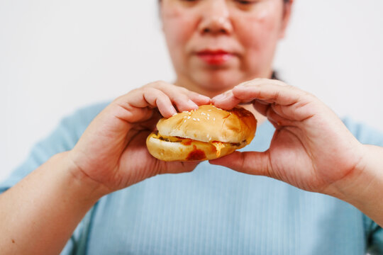 A middle-aged Asian woman with an open mouth is about to bite into a small burger or sandwich, held between her hands. The concept is about enjoying comfort food.