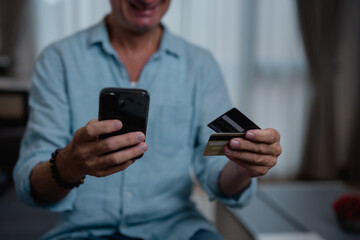 Smiling man enjoying online shopping while paying with a smartphone and credit cards, experiencing the convenience of e-commerce from the comfort of home