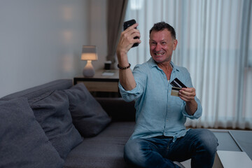 Fototapeta premium Happy mature man holding credit card and using smartphone for online shopping while sitting on sofa at home, he is taking selfie and smiling at camera