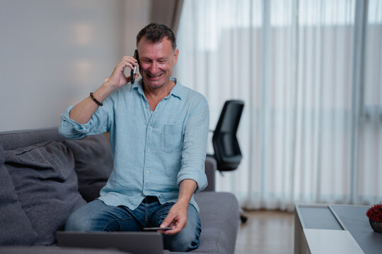 Smiling man sitting on a sofa in a bright living room, using a laptop and holding a credit card while engaged in a phone call, enjoying the convenience of online shopping