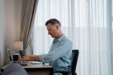Mature entrepreneur sitting at his desk in home office, using laptop and taking notes, working remotely in a quiet and comfortable environment with natural light
