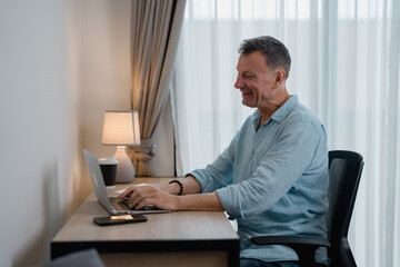 Happy businessman sitting at his desk using laptop computer working from home office enjoying online meeting, typing on keyboard, smiling, looking at screen, drinking coffee, modern interior design