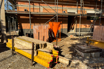 A construction site showcases various building materials, including wooden beams and bricks, surrounded by scaffolding. The orange funnel stands out against the brickwork during daytime.