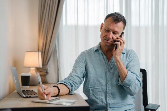 Concentrated middle-aged male entrepreneur in casual clothes having phone call conversation and writing notes in notebook while sitting at desk with laptop in home office