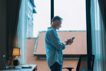 Obraz premium Businessman standing next to a window in an office, using a smartphone while a desk with a laptop and coffee cup is visible in the foreground, creating a modern work atmosphere