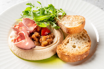 Gourmet hummus plate with mortadella slices, chickpeas, tomatoes, arugula, and toasted bread, served on a white dish with herb oil drizzle. Mediterranean appetizer presentation