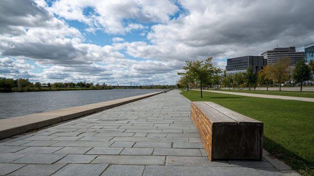 Empty park bench on paved riverside walkway.  Clouds and city in background