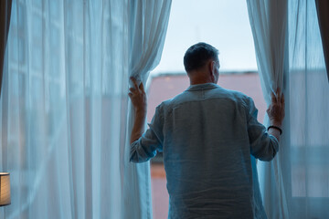 Senior man opening curtains and gazing out the window, taking in the expansive city view from his home, feeling a deep sense of loneliness and contemplation in the quiet moment