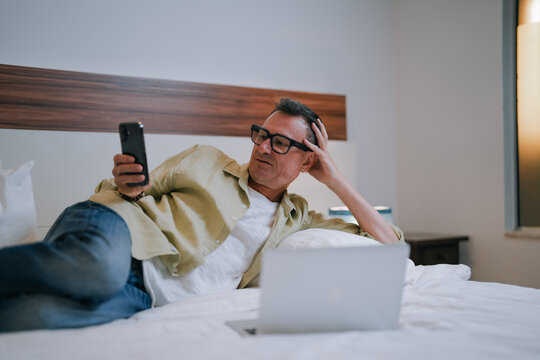 Mature businessman wearing glasses is using smartphone and laptop while lying on bed in hotel room, he is touching his head with hand looking thoughtful