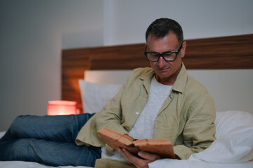 Fototapeta premium Relaxed middle-aged man wearing glasses enjoying reading a book while lying comfortably in bed at night with a soft lamp illuminating the room, creating a cozy atmosphere for bedtime reading