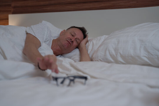 Tired senior man wearing white pajamas is resting comfortably in bed, reaching for his glasses as morning light filters into the serene bedroom, promoting a sense of relaxation and wellbeing