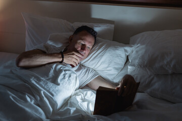 Thoughtful mature man lying in bed, reading a book under the soft glow of dim light, savoring a peaceful moment of relaxation before drifting off to sleep in his cozy bedroom