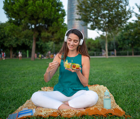 Young woman enjoying a healthy lunch break in the city park, listening music with headphones