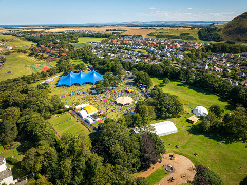 Aerial view of a vibrant festival unfolds with tents and crowds nestled among lush greenery, overshadowed by the iconic Arthur's Seat, Edinburgh, Scotland, United Kingdom.