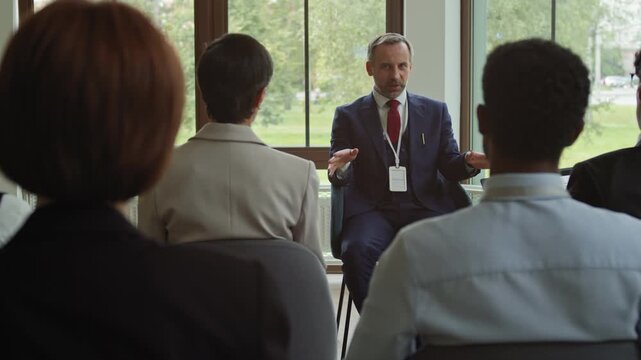 Rear view of several intercultural delegates sitting in rows and listening to confident experienced business coach in formalwear explaining main points of his report during training