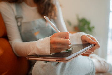Young woman artist wearing casual clothes is drawing on a digital tablet with a stylus while sitting comfortably on a sofa in her living room, working from home