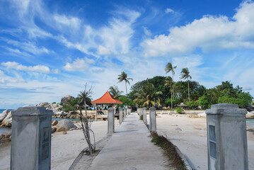A concrete walkway leads to a small island with palm trees and a gazebo, under a bright blue sky with scattered clouds.