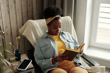 Horizontal high angle portrait shot of young African American woman wearing casual outfit relaxing in chair at home reading novel