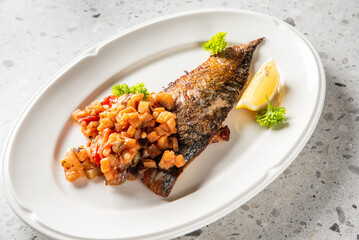 Grilled fish fillet with crispy skin served on a white plate, topped with vegetable relish, garnished with lemon wedge and parsley. Gourmet seafood dish on textured background