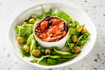 Baked eggplant dish in white ramekin topped with tomato sauce, served with arugula and green olives on a white plate. Healthy vegetarian meal, clean presentation, top view