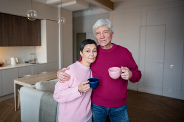Older married couple embracing and drinking coffee tea and looking at camera. Senior spouses rest together, retired life contentment, closeness affection in later years, emotional stability in old age