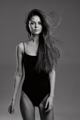 Naklejka premium Black and white portrait of a young woman with long hair wearing a black bodysuit confidently posing against a plain background.