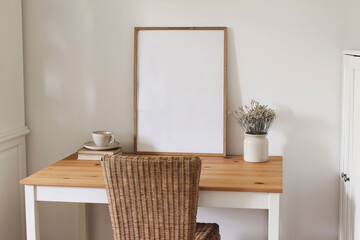 Minimalist home office interior with desk, wicker chair. Blank wooden picture frame, poster mockup, dried flowers in ceramic vase. Coffee cup on books. Bright neutral tones. Scandinavian living room