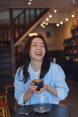 Cheerful Asian woman smiling and laughing while drinking hot coffee at modern cafe, representing freelance lifestyle, digital nomad, and online business success.