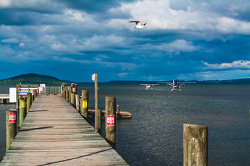 Dramatic skies over Lake Rotorua with a rustic wooden pier leading to calm waters, where floatplanes rest against a backdrop of distant hills and scenic beauty, capturing adventure and tranquility