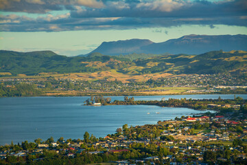 Fototapeta premium A breathtaking view of Rotorua city nestled beside Lake Rotorua, with rolling green hills and distant mountains under a dramatic sky, showcasing the harmony of nature and community