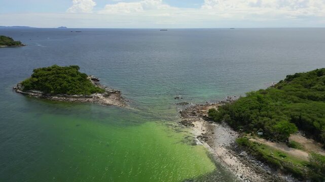 A stunning aerial drone shot captures a small, rocky island covered in green trees, surrounded by the calm, clear sea. The view also features a larger island or mainland coast with lush forests in the