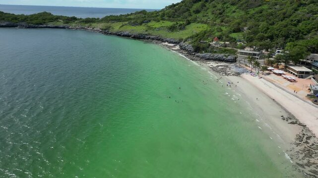 A scenic aerial drone shot capturing a beautiful tropical beach with calm, turquoise water. People are seen swimming in the sea and relaxing on the sandy shore, which is lined with small buildings and