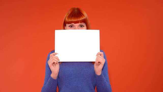 A woman with orange hair holding a blank white sheet in front of her face against an orange background