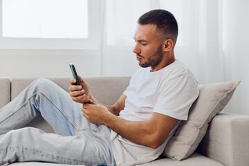 Thoughtful man using smartphone while relaxing on a cozy couch, featuring natural light and soft tones for a modern living space ambiance.