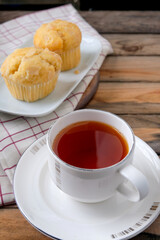 Hot cup of tea served on white saucer with freshly baked almond muffins in background on rustic wooden table, cozy breakfast or snack scene ideal for cafe, bakery, and lifestyle concept.