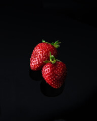 Close-up of two fresh strawberries with green stems on a reflective black surface. The red strawberries stand out against the dark background, highlighting their natural texture and vibrant color.