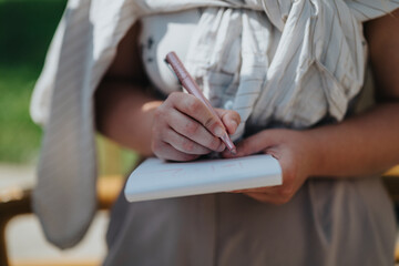 Captured moment of an student writing notes on a small notepad outdoors. The scene feels casual and focused, emphasizing creativity and writing. The sunlight enhances the overall uplifting ambience.