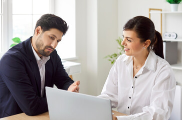 Obraz premium Two confident business people wearing formal clothing looking at laptop monitor screen at workplace discussing company finance sitting at the desk. Employees and coworkers working in office.