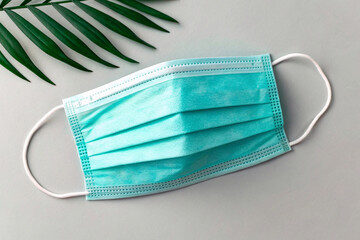 Turquoise disposable medical face mask with white elastic ear loops on neutral background with green palm leaf decoration above.
