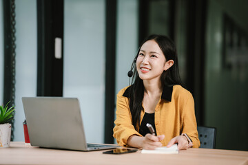 A cheerful young woman wearing a headset communicates through her laptop in a modern office, providing customer support or engaging in an online business meeting with confidence.
