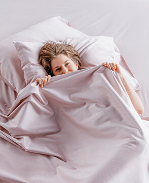 A cheerful woman lies comfortably in a bed surrounded by soft silk sheets. She peeks out from under the covers, enjoying a cozy morning at home filled with tranquility.