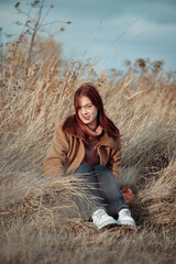 Smiling teenage girl with red hair in a warm coat, seated in tall dry grass, enjoying an autumn day.