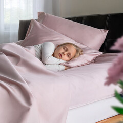 A young child is peacefully sleeping wrapped in soft pink bedding on a cozy bed. The morning light softly illuminates the room, creating a serene atmosphere.