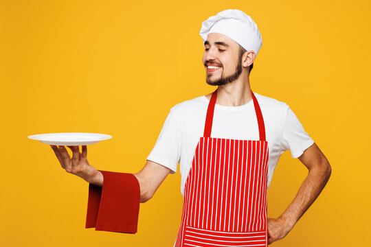 Young smiling happy housewife housekeeper chef cook baker man wear red apron toque hat hold in hand empty plate napkin isolated on plain yellow orange background studio portrait. Cooking food concept.
