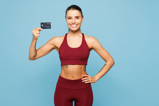Young fitness trainer instructor sporty woman sportsman wear red sportswear training in home gym hold hand mock up of credit bank card isolated on plain blue background. Workout sport fit abs concept.