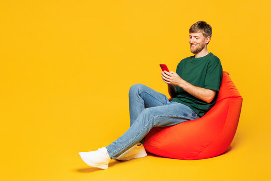 Full body young smiling happy man he wear green t-shirt casual clothes sit in bag chair hold in hand use mobile cell phone isolated on plain yellow orange background studio portrait Lifestyle concept - Powered by Adobe