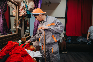 A young actor in a grey jumpsuit attends to garments on a table, surrounded by costumes and props in a backstage area of a theater. © qunica.com